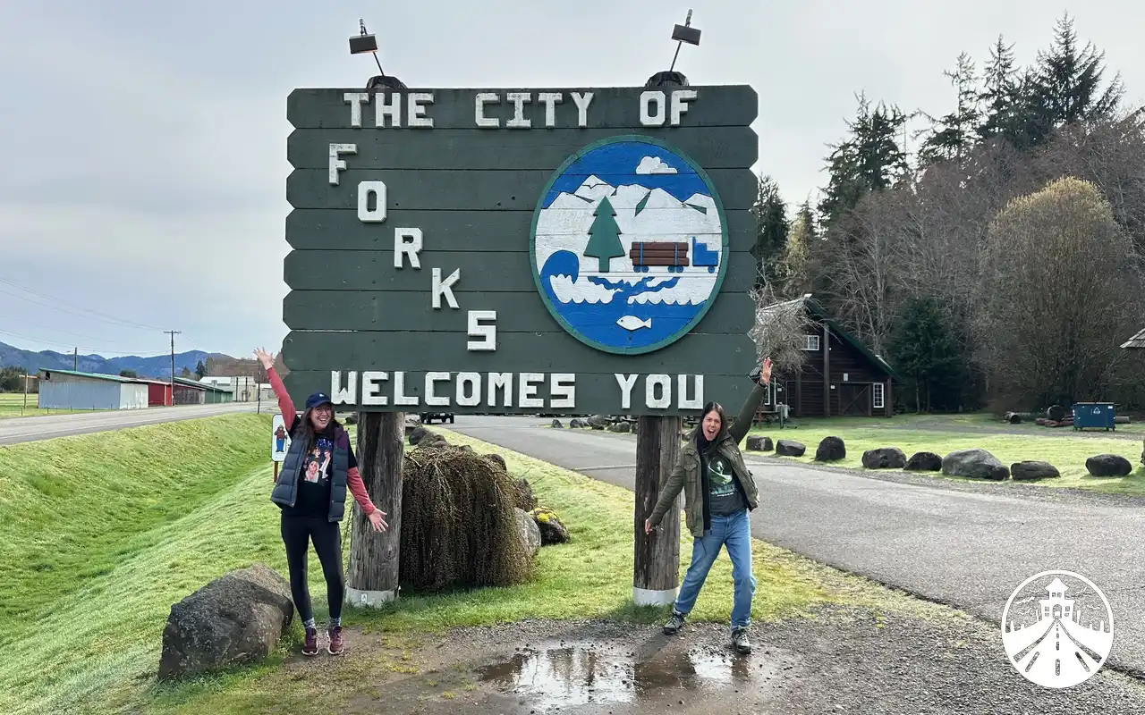 Gem and Glenna standing in front of the “Welcome to Forks” sign on a cloudy day in Washington.
